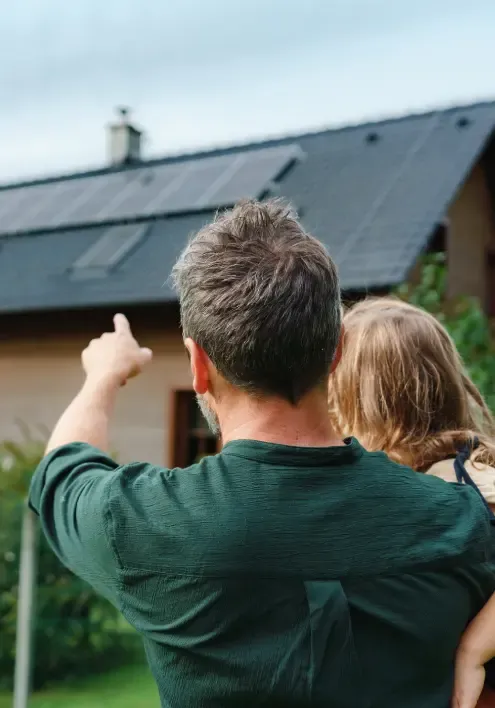 Far med barn på armen som peker opp mot solcellepanelene på taket av huset