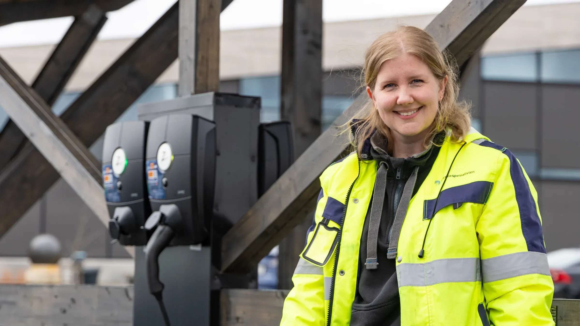 Smilende ansatt i refleksjakke ved siden av ladestasjoner