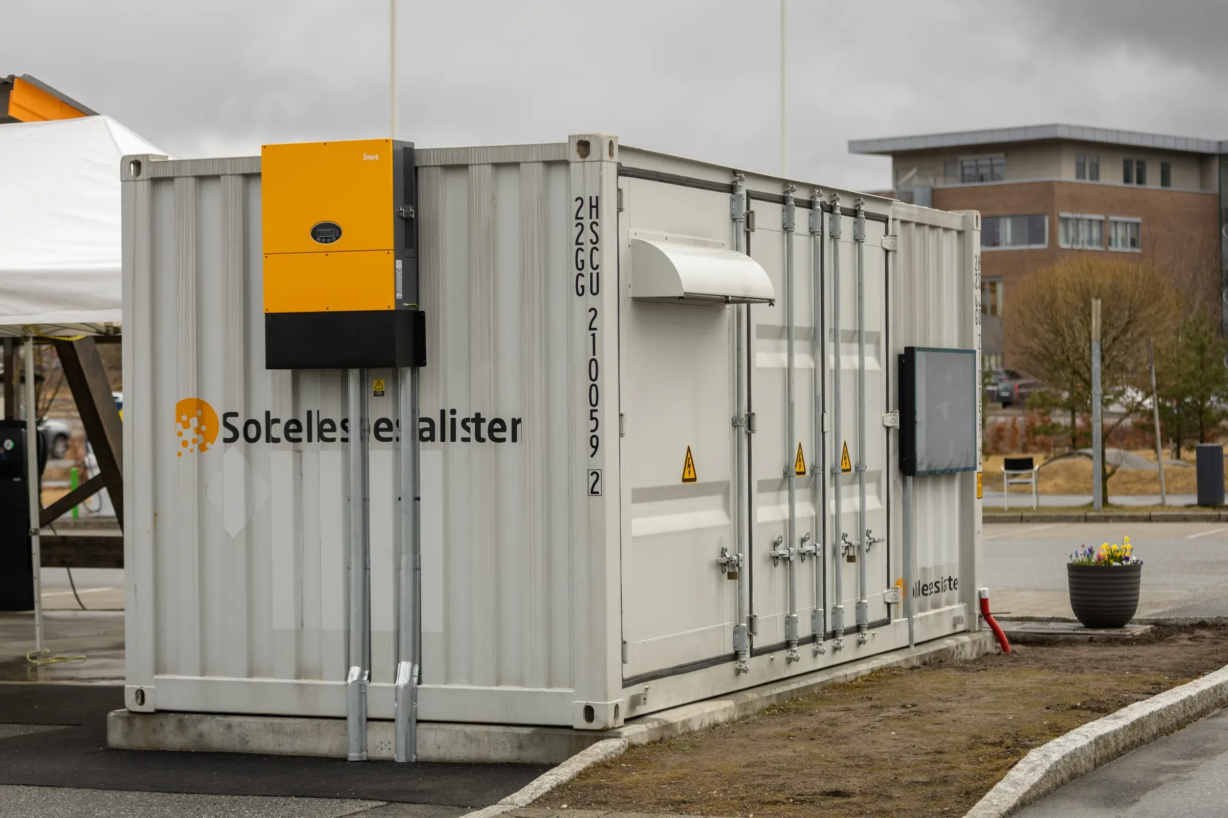 Hvit batterikontainer med en oransje vekselretter montert på endeveggen, plassert utendørs ved et næringsbygg