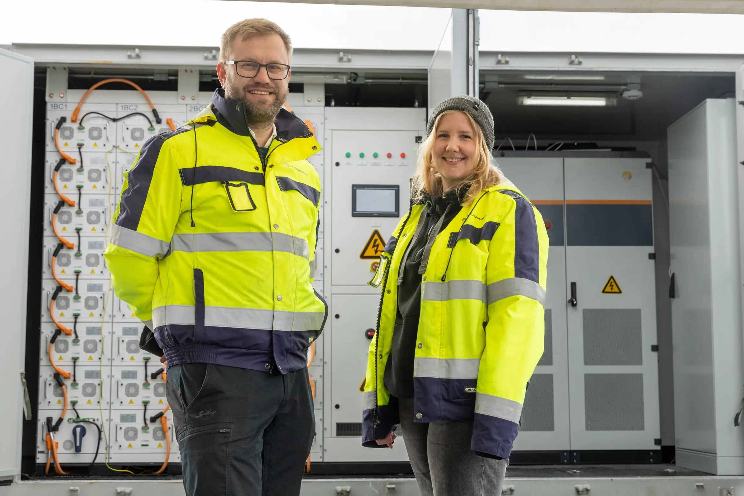 To ansatte i refleksjakke foran et åpent batterianlegg med batterimoduler og styreskap