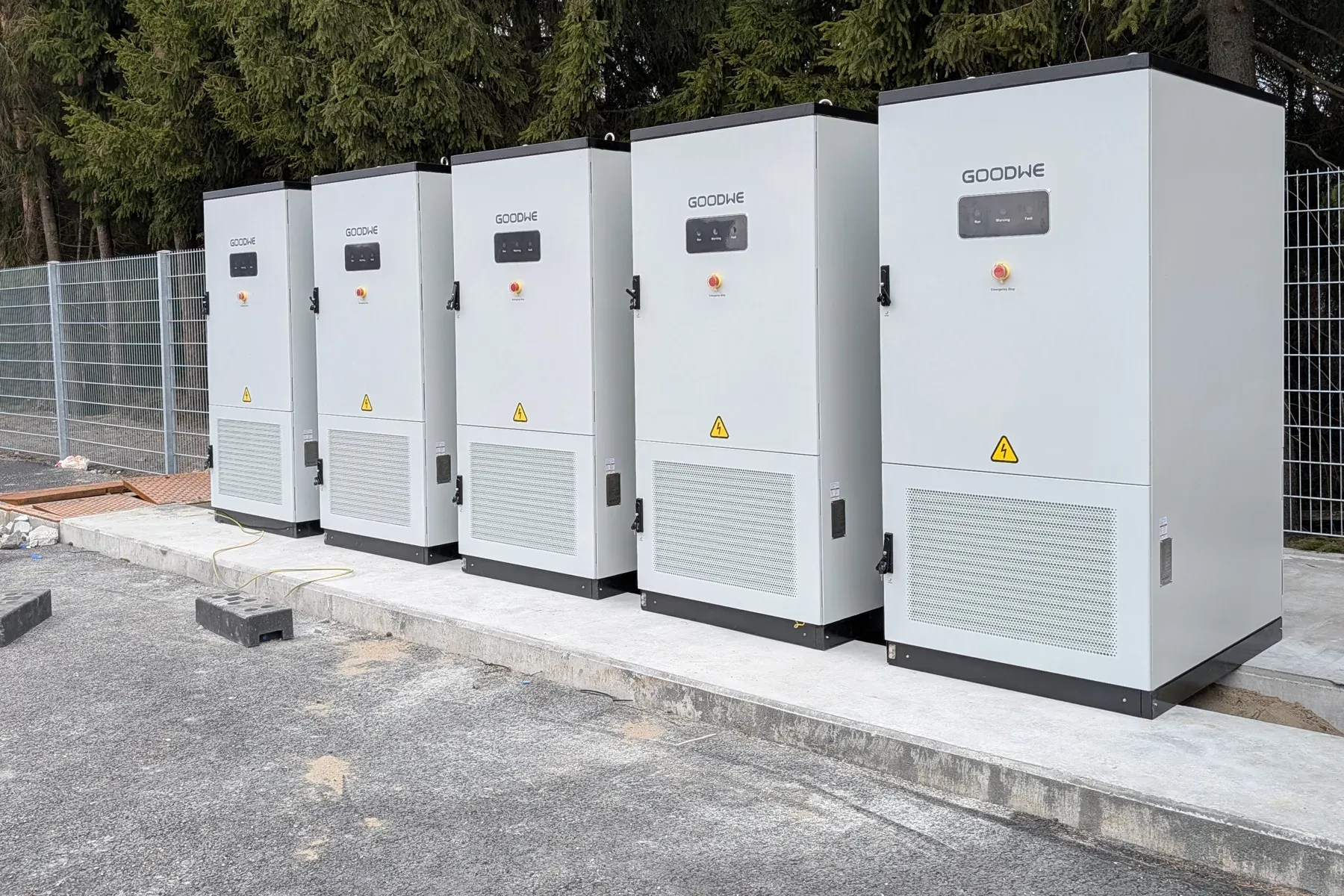 Fem GoodWe-batterikabinetter på rekke på et betongfundament utendørs, med granskog bak