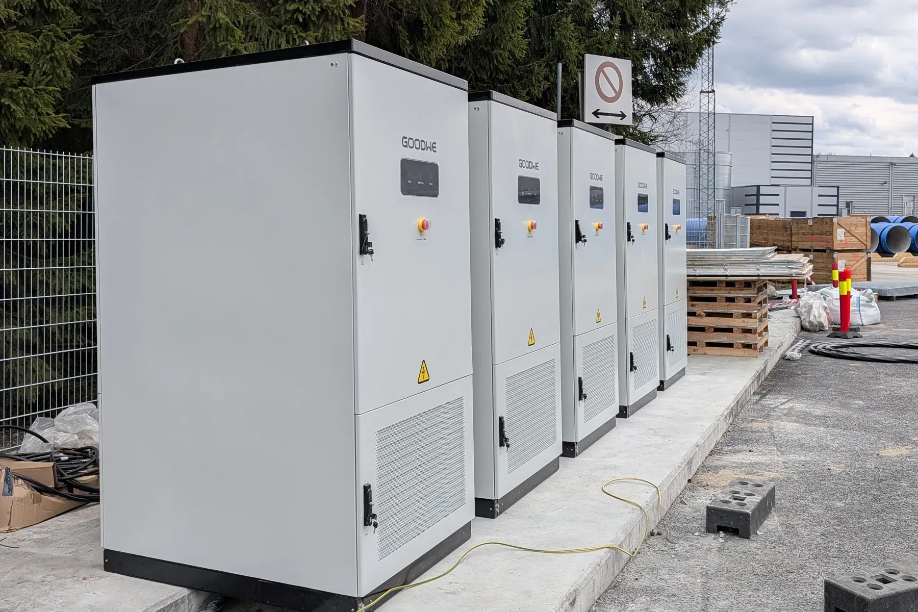Fem batterikabinetter på rekke på et betongfundament utendørs, med et forbudsskilt og industribygg i bakgrunnen
