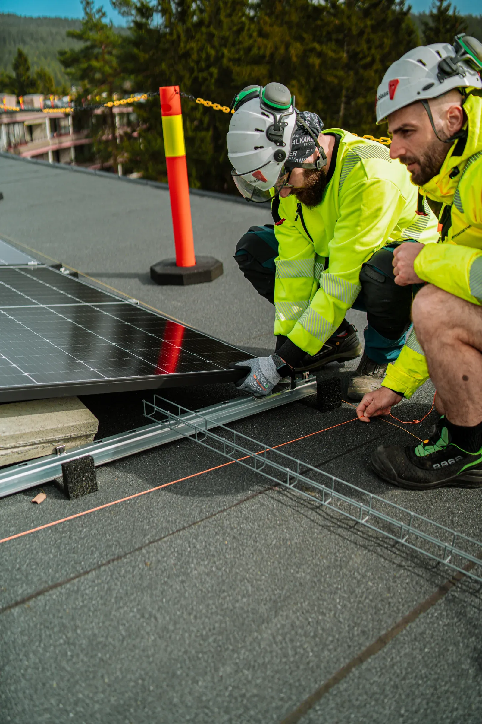 To montører med hjelm og refleksjakke fester et solcellepanel på et flatt tak, med en sikkerhetskjegle bak