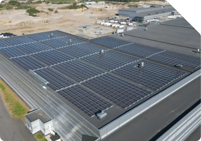 Dronefoto av et stort flatt næringstak som er dekket av solcellepaneler i lange, lavt hellende rader, med et industriområde i bakgrunnen