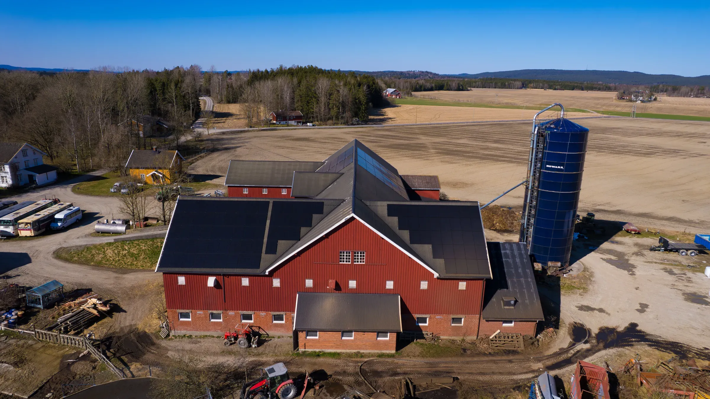 Rød driftsbygning med solcellepaneler på begge takflatene, med silo og jorder rundt, sett fra lufta