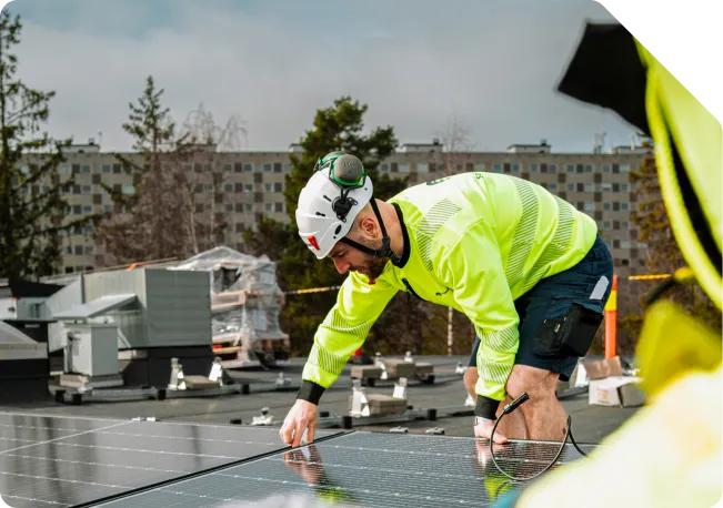 Montør i gul arbeidsjakke og hvit hjelm som legger DC-kabel mellom solcellepaneler på et flatt næringstak